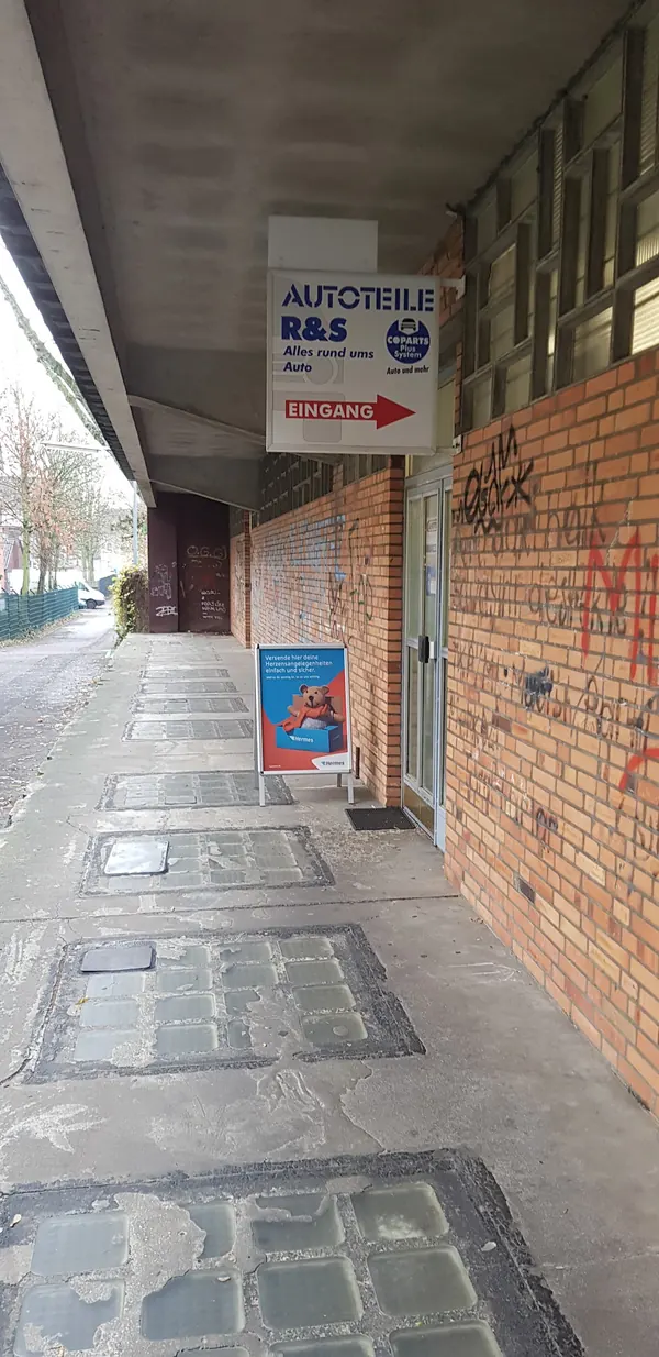 Weg am Gebäude entlang mit Hinweisschild „Autoteile R&S – Eingang“